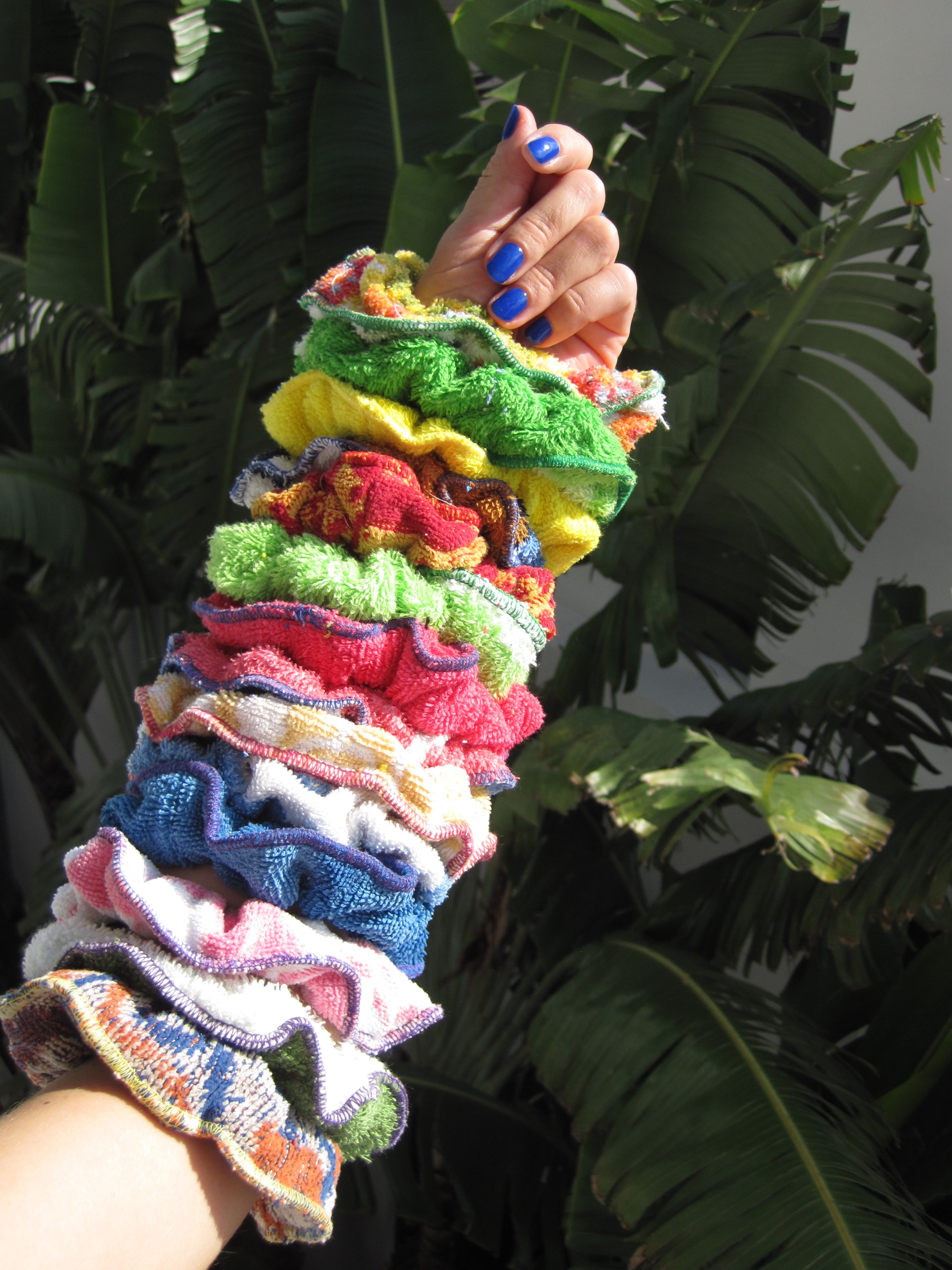 Arm adorned with multiple colorful fabric scrunchies, raised against a backdrop of tropical green leaves.