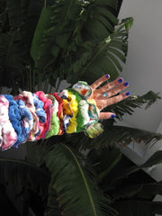 Extended arm wearing multiple colorful fabric scrunchies, with blue-painted nails and a ring, set against tropical green leaves.